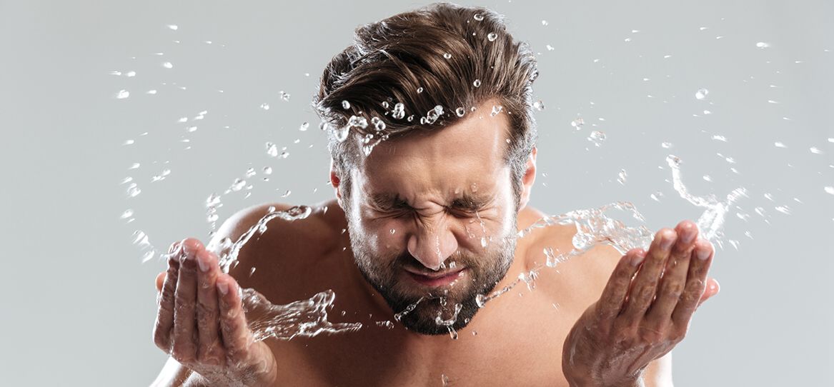 Junger Mann mit kurzem Vollbart vor neutralem grauen Hintergrund spritzt sich mit beiden Händen Wasser ins Gesicht