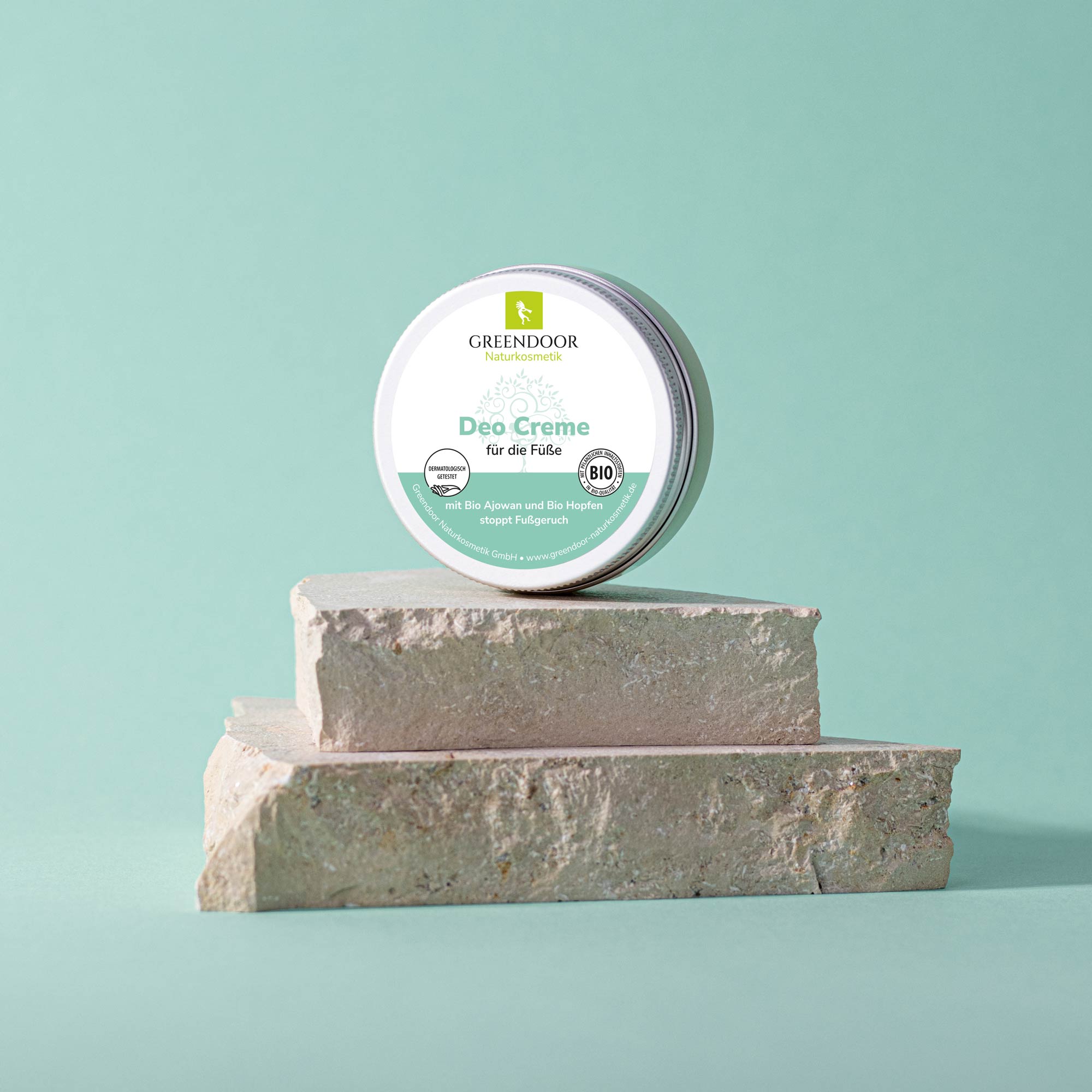 Greendoor Deo Creme für die Füße - Bio Ajowan und Hopfen, stoppt Fußgeruch Eine Dose Greendoor Deo Creme für die Füße steht auf einem Steinpodest vor einem mintgrünen Hintergrund